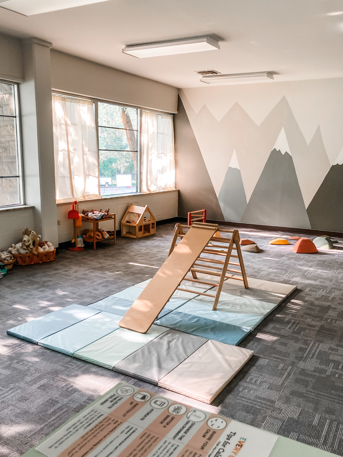 Sunlit classroom with soft play mats, a wooden climbing ramp and a painted mountain mural