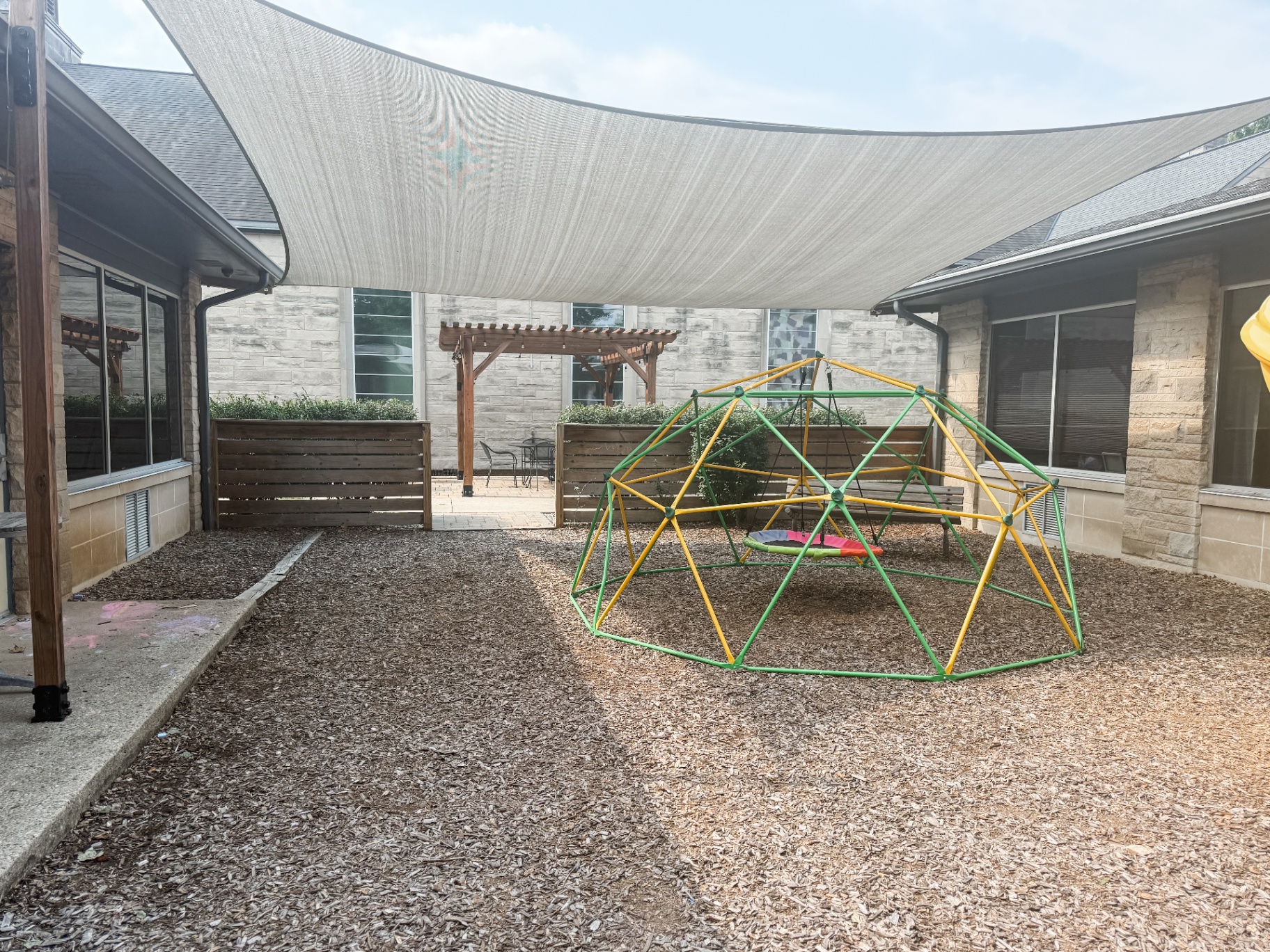 Playground with a large shade sail stretched over the courtyard and a pergola seating area beyond