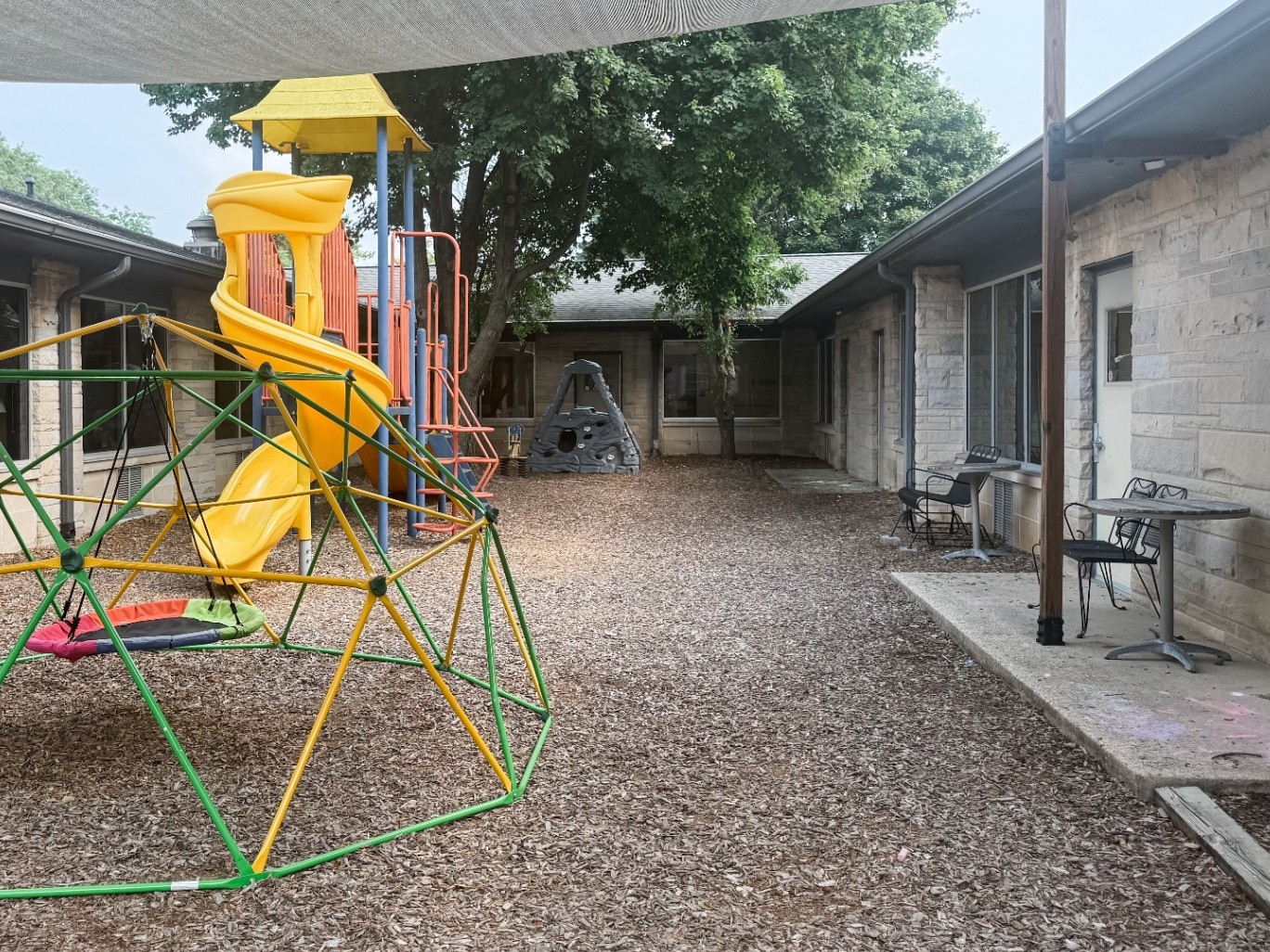Courtyard playground with a yellow spiral slide and a green climbing dome under a shade sail