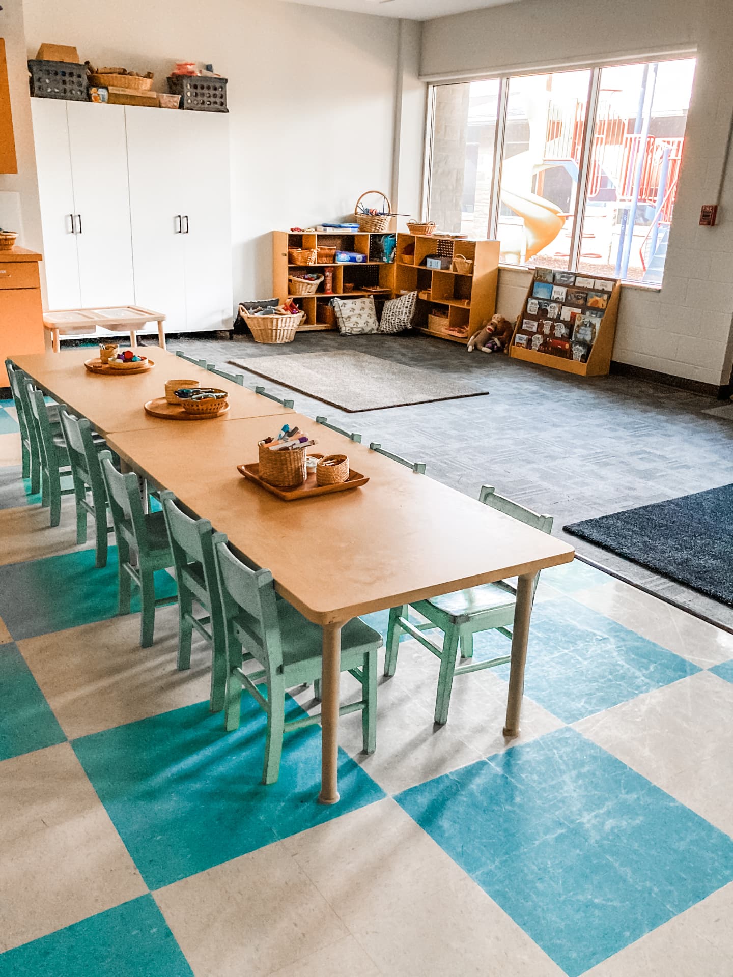 Sunlit classroom with a long wooden table, green chairs and a view of the playground