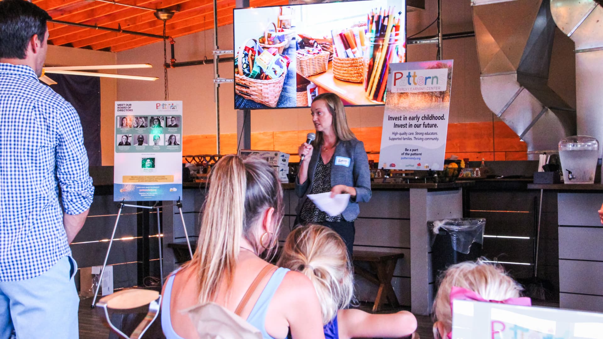 A board member speaking into a microphone beside Patterns signage and a slideshow